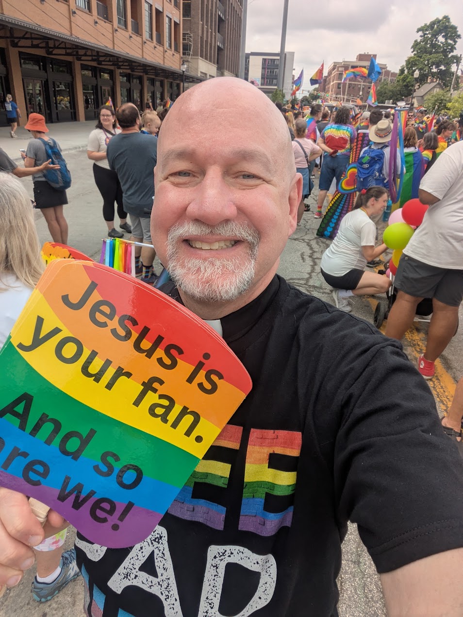 Shawn at Pride holding a rainbow sign that reads 'Jesus is your fan. And so are we!'